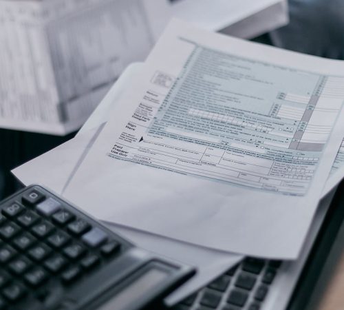 A young adult using a calculator and laptop to manage finances at home, surrounded by papers.